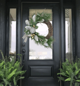Black front door with a pine and silver holiday wreath, flanked by two large fern plants in black pots, and a plain doormat on the porch. - Spruced Studio Upscale Workshop