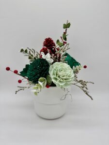 A white pot holds an arrangement of artificial flowers, red berries, pine cones, and green leaves against a plain white background. - Spruced Studio Upscale Workshop