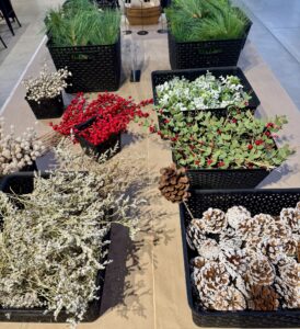 Black baskets on a table filled with pine needles, red berries, white dried flowers, green leaves, and snow-dusted pinecones, arranged for craft or decoration purposes. - Spruced Studio Upscale Workshop