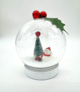 Clear ornament shaped like a snow globe, containing a small snowman, a pine tree, artificial snow, two red berries, and a green leaf on top. - Spruced Studio Upscale Workshop