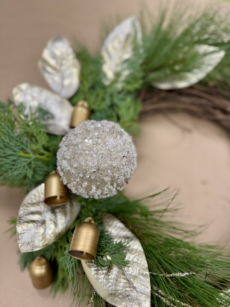 A holiday wreath decorated with gold leaves, gold bells, green pine branches, and a large glittery silver ornament. - Spruced Studio Upscale Workshop