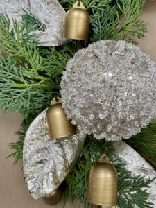 A close-up of a festive arrangement featuring green pine branches, frosted silver leaves, a textured silver ornament, and small gold bells. - Spruced Studio Upscale Workshop