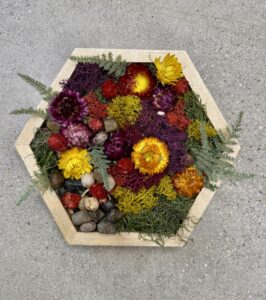 A hexagonal wooden tray filled with colorful dried flowers, green moss, small rocks, and sprigs of fern, placed on a gray concrete surface. - Spruced Studio Upscale Workshop