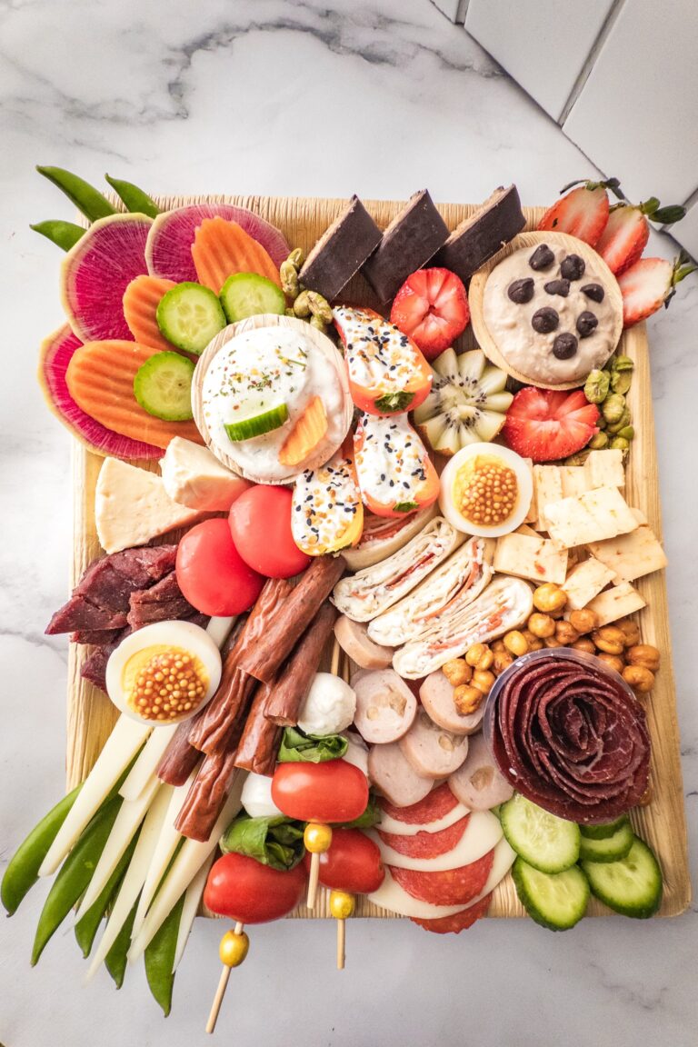 A charcuterie board with assorted meats, cheeses, crackers, vegetables, fruit, chocolate, dip, and garnishes, arranged on a square wooden tray. - Spruced Studio Upscale Workshop
