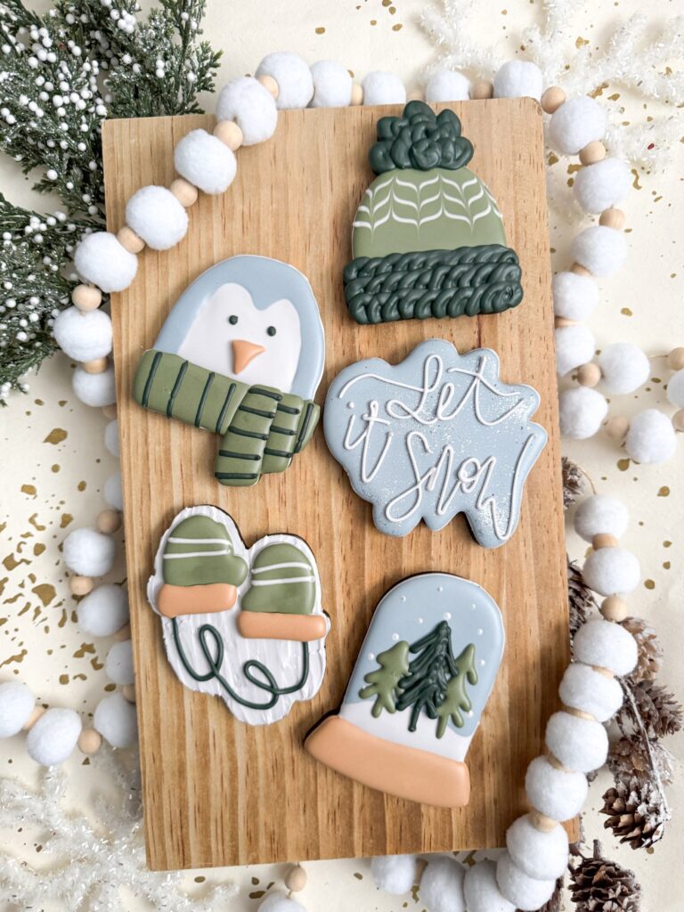 A wooden board displays five decorated winter-themed cookies: a penguin, a knitted hat, “let it snow” text, mittens, and a tree in a snow globe. White garland and winter decor surround the board. - Spruced Studio Upscale Workshop