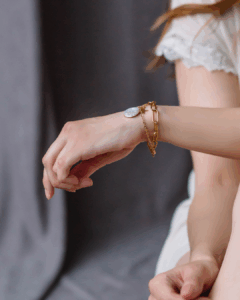 A person wearing a white short-sleeved top displays a gold chain bracelet with a round pearl charm on their wrist. - Spruced Studio Upscale Workshop