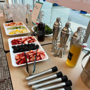 A table set with sliced fruits, bottles, cocktail shakers, tongs, shot glasses, and plastic cups, prepared for a beverage or cocktail station. - Spruced Studio Upscale Workshop