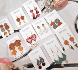 A display of ten pairs of colorful, uniquely designed earrings on branded cards, arranged neatly on a fabric surface with flowers as decoration. - Spruced Studio Upscale Workshop