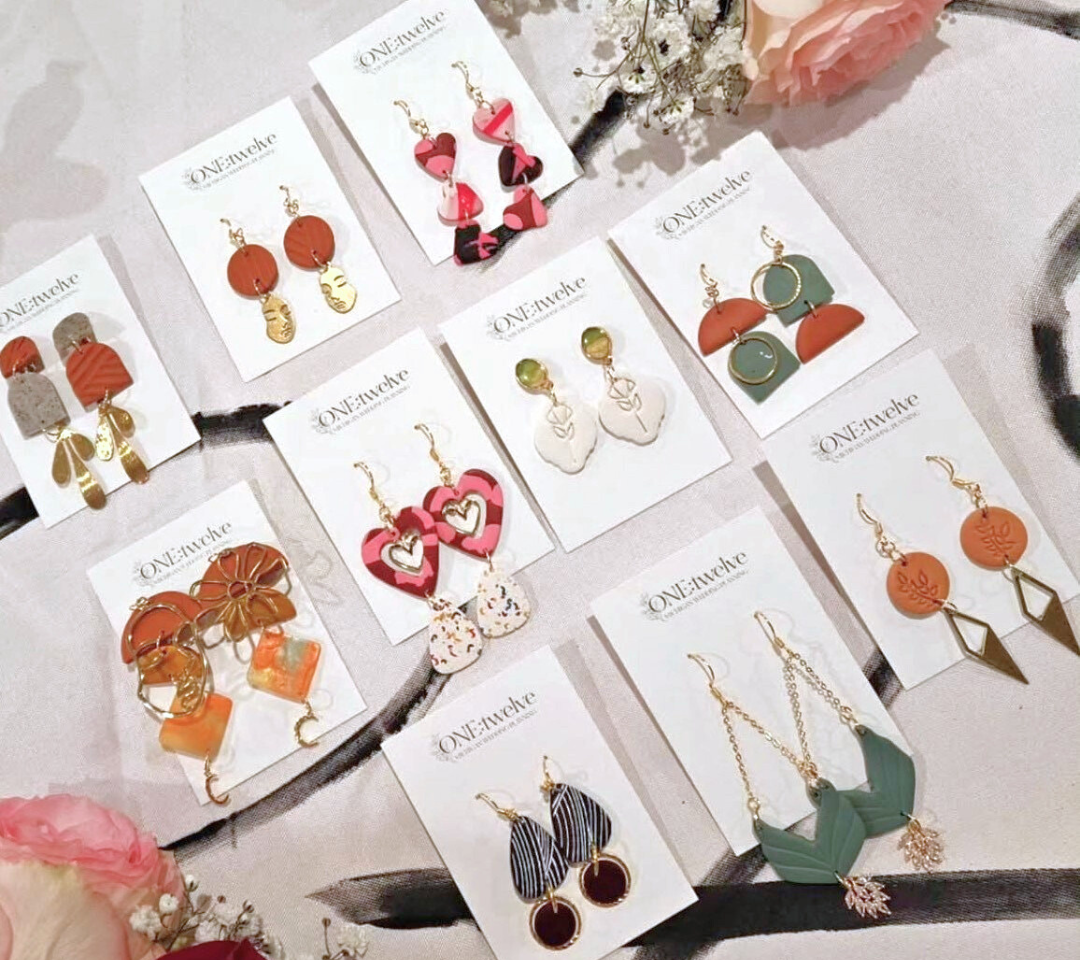 A display of ten pairs of colorful, uniquely designed earrings on branded cards, arranged neatly on a fabric surface with flowers as decoration. - Spruced Studio Upscale Workshop