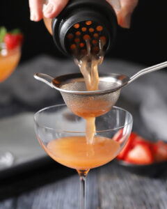 A hand pours a cocktail through a strainer into a stemmed glass, with sliced strawberries blurred in the background. - Spruced Studio Upscale Workshop