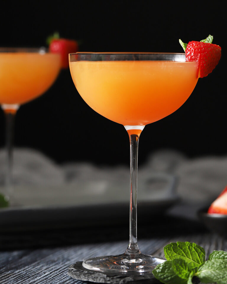 A stemmed glass filled with an orange cocktail garnished with a fresh strawberry on the rim, with another similar drink blurred in the background. - Spruced Studio Upscale Workshop