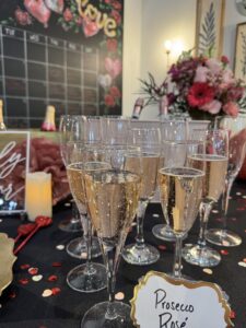 Several glasses of sparkling rosé wine are arranged on a table decorated with confetti, flowers, and a sign reading "Prosecco Rosé." A "Love" sign and bottles are visible in the background. - Spruced Studio Upscale Workshop