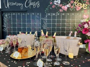 Table with assorted champagne glasses, two ice buckets filled with bottles, and a floral centerpiece; a chalkboard calendar for February and March is in the background. - Spruced Studio Upscale Workshop