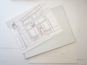 A hand-drawn line art sketch of a storefront sits on top of two sheets of blank or lightly sketched paper on a white surface. - Spruced Studio Upscale Workshop