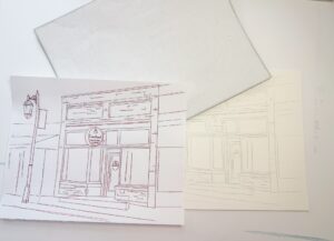 A sheet of tracing paper, a line drawing of a storefront, and a faint transferred outline of the same drawing on another sheet of paper are shown on a white surface. - Spruced Studio Upscale Workshop