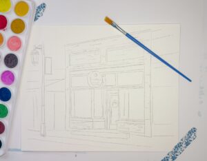A sketched outline of a storefront on watercolor paper with a paintbrush and a watercolor palette placed beside it. - Spruced Studio Upscale Workshop