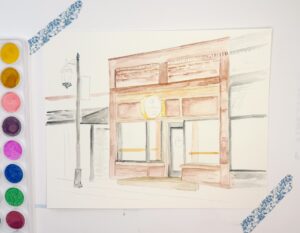 A watercolor painting of a storefront in progress, with some areas detailed and others unfinished, sits on a white surface beside a watercolor palette. - Spruced Studio Upscale Workshop