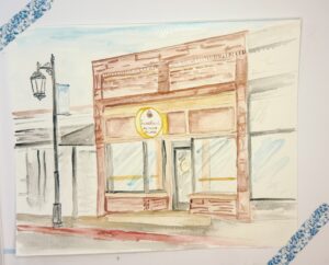 Watercolor painting of a brick storefront with a round yellow sign reading "Sweetheart Bakery & Cafe," flanked by large windows and a streetlamp on the left. - Spruced Studio Upscale Workshop