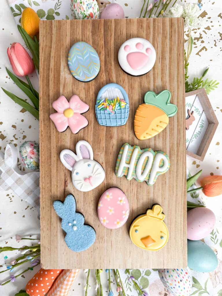 Nine decorated Easter-themed sugar cookies, including eggs, a bunny, a chick, a carrot, a basket, a flower, and the word "HOP," arranged on a wooden board with spring decorations surrounding them. - Spruced Studio Upscale Workshop