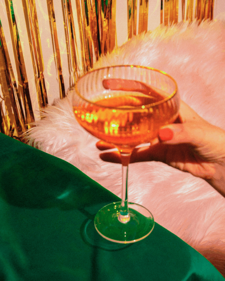 A hand with painted nails holds a coupe glass filled with a golden drink, next to a green tablecloth and a pink faux fur surface, with gold tinsel in the background. - Spruced Studio Upscale Workshop