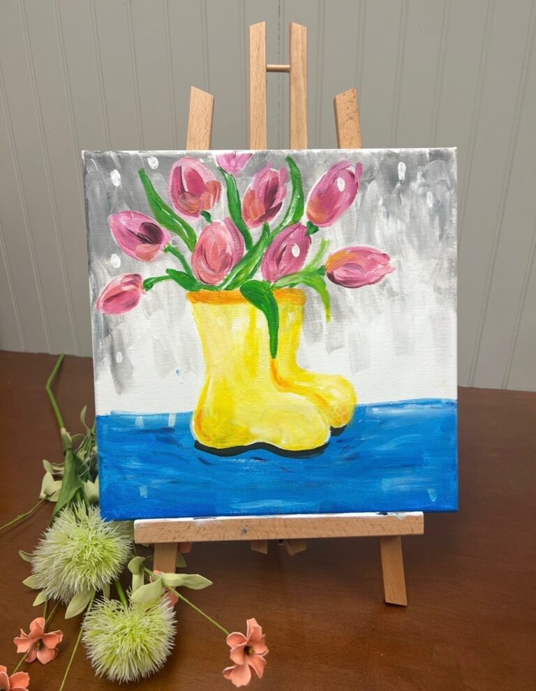 A painting of pink tulips arranged in yellow rain boots is displayed on a wooden easel, with artificial flowers placed nearby on a brown table. - Spruced Studio Upscale Workshop