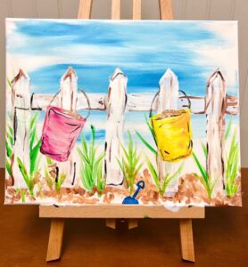 A painting on an easel depicts a white fence with a pink and a yellow bucket hanging on it, green grass, brown sand, and a blue sky in the background. - Spruced Studio Upscale Workshop