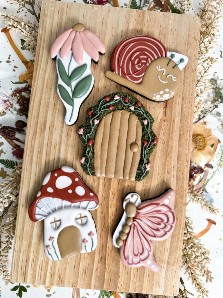 Five decorated cookies shaped as a pink flower, a smiling snail, a round wooden door, a mushroom house, and a butterfly on a wooden board. - Spruced Studio Upscale Workshop