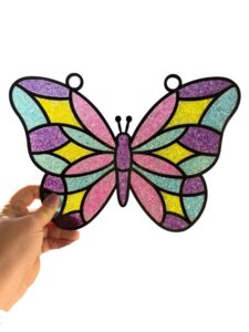 A hand holds a butterfly-shaped stained glass ornament with colorful sections in pink, yellow, teal, and purple, outlined in black. - Spruced Studio Upscale Workshop