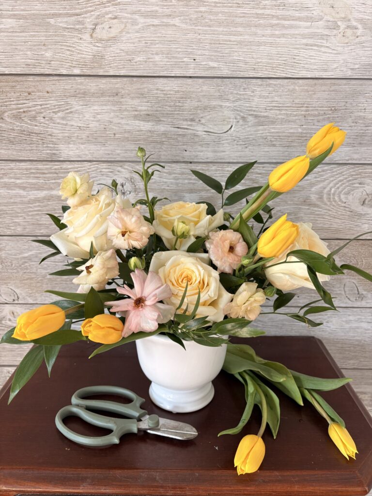 A white vase with yellow tulips, white roses, and greenery sits on a wooden table next to a pair of green-handled floral scissors. - Spruced Studio Upscale Workshop