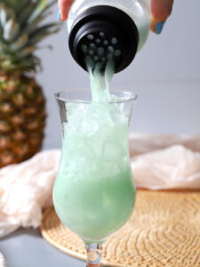 A light green drink is being poured from a shaker into a glass filled with ice, with a pineapple and woven mat in the background. - Spruced Studio Upscale Workshop