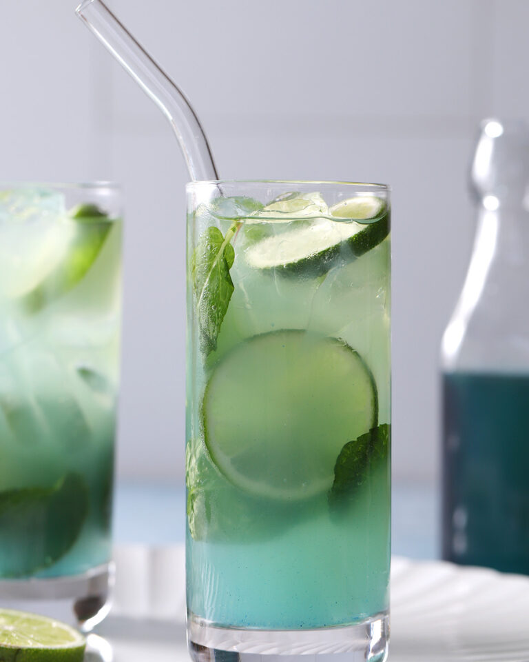 A tall glass of blue-green iced drink with lime slices, mint leaves, and a clear straw, set on a light surface with another similar drink and a bottle in the background. - Spruced Studio Upscale Workshop