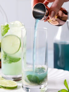 A hand pours blue liquid from a jigger into a glass with mint leaves; a drink with lime and mint, a bottle, and lime slices are visible in the background. - Spruced Studio Upscale Workshop