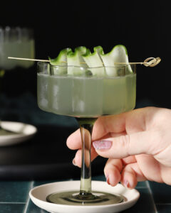 A hand holds a coupe glass filled with a green cocktail, garnished with ribboned cucumber slices on a skewer, placed on a small white plate. - Spruced Studio Upscale Workshop