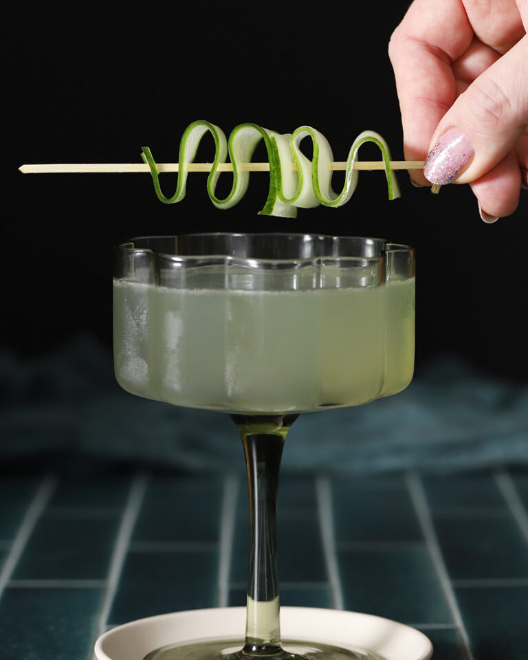 A glass of light green cocktail on a saucer, with a hand holding a skewer of spiral-cut cucumber above the drink. - Spruced Studio Upscale Workshop
