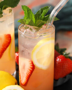 A glass of iced lemonade with strawberry slices, a lemon wheel, fresh mint garnish, and a clear straw. - Spruced Studio Upscale Workshop