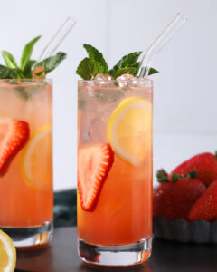 Two tall glasses of iced strawberry lemonade garnished with lemon slices, strawberry slices, mint leaves, and clear straws. A small pile of strawberries is in the background. - Spruced Studio Upscale Workshop