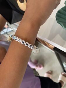 A person wears a bracelet with white beads spelling "SUMMER," a gold star bead, pastel beads, and a silver peace sign charm. - Spruced Studio Upscale Workshop