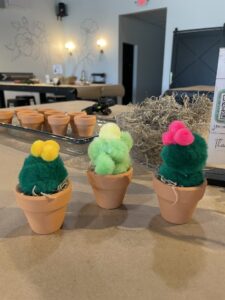 Three small artificial cacti made from green pom-poms and colorful pom-pom tops are displayed in mini clay pots on a table. - Spruced Studio Upscale Workshop