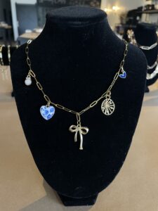 Gold chain necklace with a bow pendant, blue heart charm, round white charm, blue circle charm, and a round gold charm, displayed on a black stand. - Spruced Studio Upscale Workshop