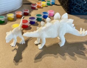 Two white plastic dinosaur figures, one larger and one smaller, are placed on a table next to a set of small paint pots in various colors. - Spruced Studio Upscale Workshop