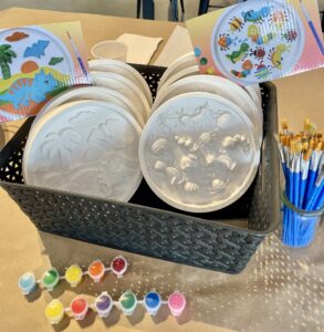 A basket holds unpainted plaster plates with dinosaur and sea life designs, paintbrushes in a jar, paint pots, and example cards showing painted versions of the designs. - Spruced Studio Upscale Workshop