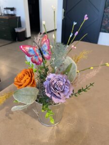 A small metal bucket holds artificial flowers, including an orange and a purple rose, assorted greenery, and a decorative butterfly on a stick. - Spruced Studio Upscale Workshop