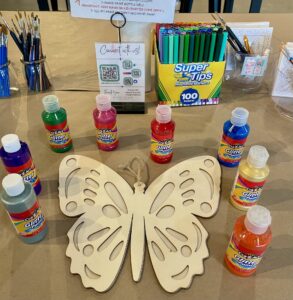 A wooden butterfly cutout is surrounded by bottles of glitter glue, Crayola Super Tips markers, paintbrushes, and a sign with painting instructions on a craft table. - Spruced Studio Upscale Workshop