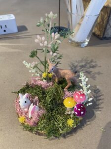 A small nature diorama featuring two white bunny figurines, a deer figurine, artificial moss, flowers, and three colorful mushroom models arranged on a round base. - Spruced Studio Upscale Workshop