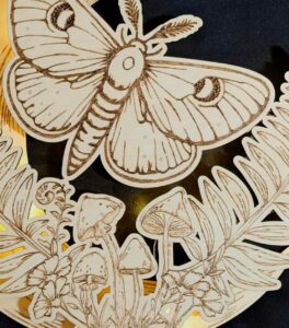 Wooden engraving of a moth with outstretched wings above mushrooms, leaves, and flowers, all intricately detailed and set against a dark background. - Spruced Studio Upscale Workshop