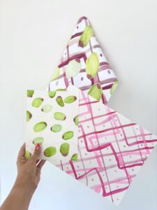 A hand holds two patterned papers, one with green watercolor shapes and one with pink grid lines; a matching pink and green patterned cloth hangs in the background. - Spruced Studio Upscale Workshop