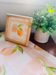 Framed peach artwork and a potted green plant sit on a table covered with a peach-patterned cloth. - Spruced Studio Upscale Workshop