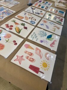 Laminated sheets with colorful watercolor illustrations of food, sea creatures, plants, and summer items arranged on a brown table. - Spruced Studio Upscale Workshop