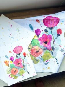 Two watercolor paintings of red and pink flowers on white paper, with one painting splattered in the background and the other on a paper towel, all placed on a wooden surface. - Spruced Studio Upscale Workshop