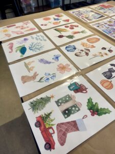 Several sheets of paper with colorful watercolor illustrations, including sea creatures, flowers, animals, and holiday-themed images, are arranged on a table. - Spruced Studio Upscale Workshop